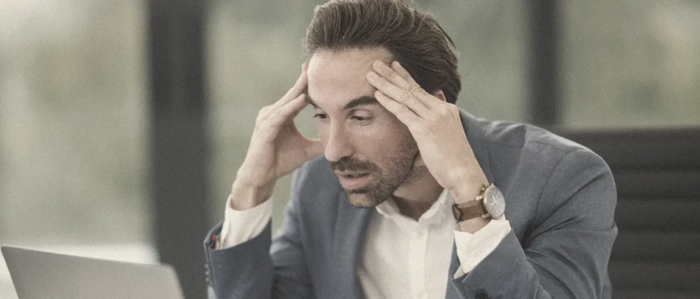 A worried man at his desk holding his head next to a laptop